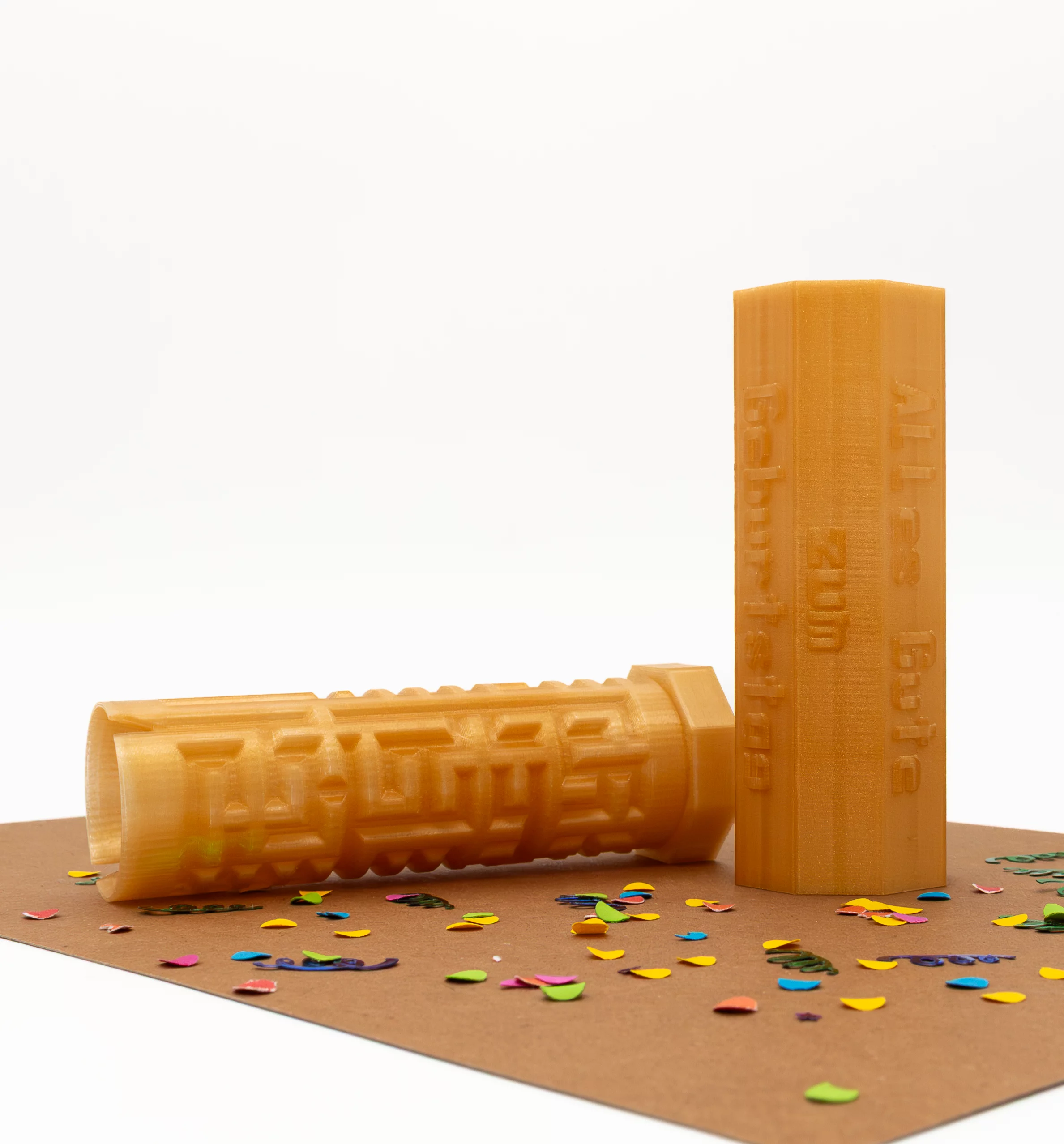 Two gold-colored 3D-printed plastic cylinders. One features the text “Happy Birthday,” the other displays a labyrinth pattern. Colorful confetti lies scattered on a brown surface.